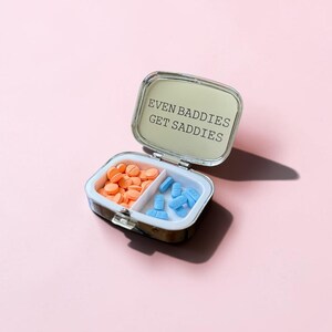 May include: A small, rectangular, silver-toned metal pill case with a hinged lid. The interior mirror features the black text "EVEN BADDIES GET SADDIES." The case is divided into two compartments, one containing round orange pills and the other blue pills.