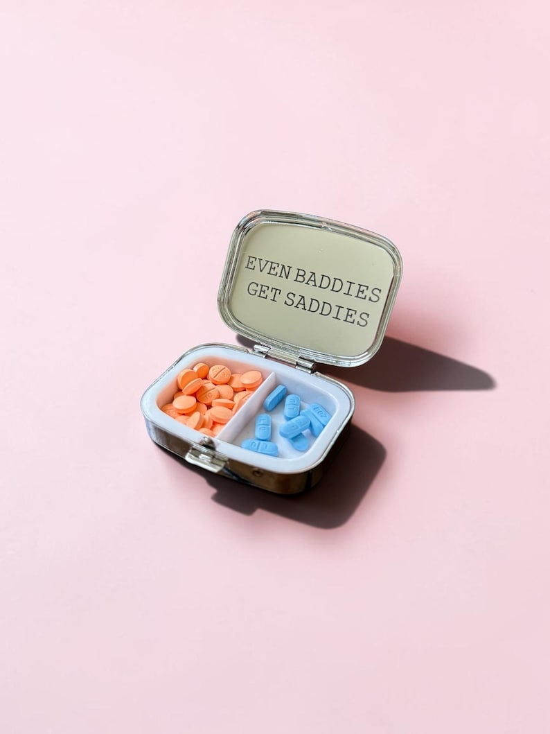 May include: A small, rectangular, silver-toned metal pill case with a hinged lid. The interior mirror features the black text "EVEN BADDIES GET SADDIES." The case is divided into two compartments, one containing round orange pills and the other blue pills.