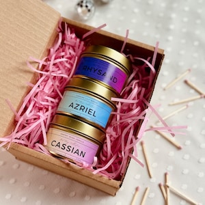 May include: A set of three gold candle tins with pink, blue, and purple ombre labels. The labels have the names "Rhysand", "Azriel", and "Cassian" on them. The candles are in a brown cardboard box with pink shredded paper.