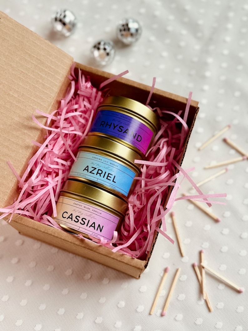 May include: A set of three gold candle tins with pink, blue, and purple ombre labels. The labels have the names "Rhysand", "Azriel", and "Cassian" on them. The candles are in a brown cardboard box with pink shredded paper.