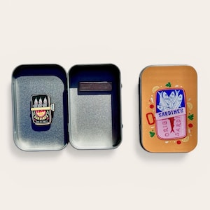 May include: A rectangular metal tin box with a hinged lid, featuring a graphic of a sardine can on the exterior. Inside the open tin, a small sardine can-shaped enamel pin is attached, displaying the text SARDINES and POUR LES TOUTTS.