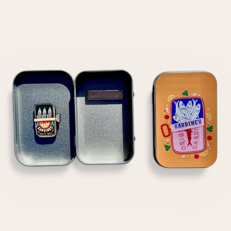 May include: A rectangular metal tin box with a hinged lid, featuring a graphic of a sardine can on the exterior. Inside the open tin, a small sardine can-shaped enamel pin is attached, displaying the text SARDINES and POUR LES TOUTTS.
