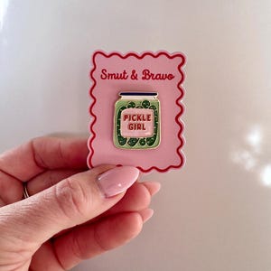 May include: A rectangular pink backing card with a scalloped red border features the text Smut & Bravo at the top. Attached to the card is a metal enamel pin shaped like a glass jar filled with green pickles, with a label reading PICKLE GIRL.