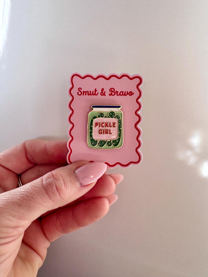 May include: A rectangular pink backing card with a scalloped red border features the text Smut & Bravo at the top. Attached to the card is a metal enamel pin shaped like a glass jar filled with green pickles, with a label reading PICKLE GIRL.