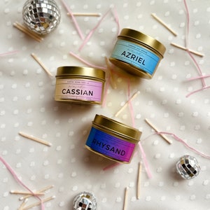 May include: Three gold candle tins with different colored labels. The labels have the names "Cassian", "Azriel", and "Rhysand" on them. The candles are surrounded by pink ribbon and wooden matches.