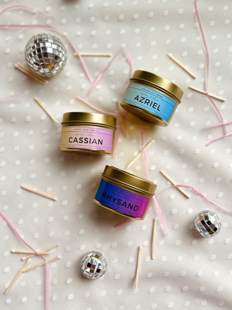 May include: Three gold candle tins with different colored labels. The labels have the names "Cassian", "Azriel", and "Rhysand" on them. The candles are surrounded by pink ribbon and wooden matches.