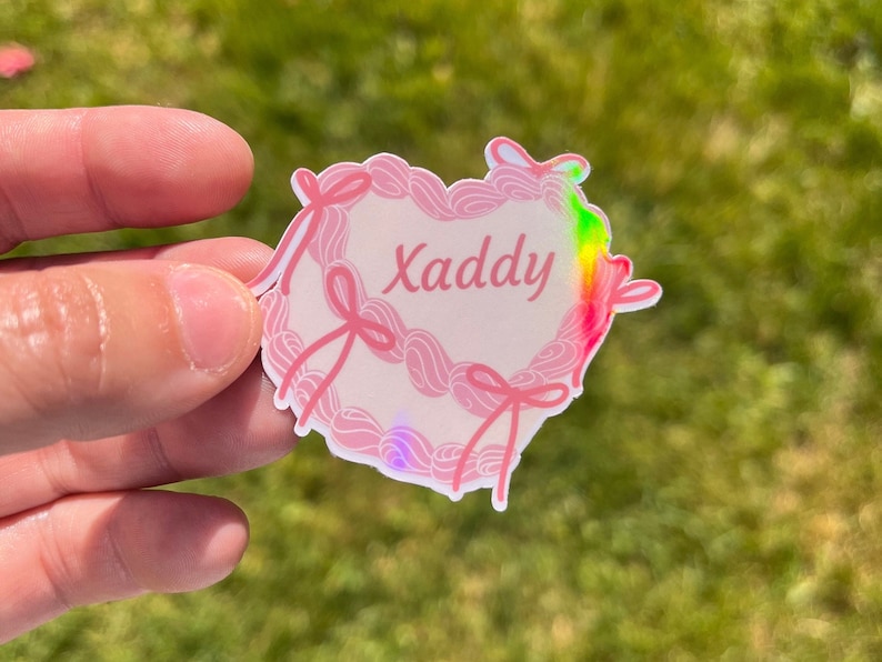 May include: A pink heart-shaped sticker with a white background and the word "Xaddy" written in black. The sticker has a holographic finish that shimmers in rainbow colors.