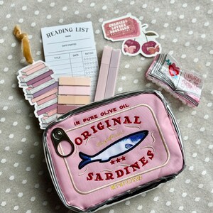 Puede incluir: Una bolsa de tela rosa con forma de lata de sardinas con el texto "Sardines Originales" y "En Aceite de Oliva Puro" bordado en ella. La bolsa tiene una cremallera plateada y una tirador plateado. La bolsa está sobre una superficie de lunares blancos y grises.