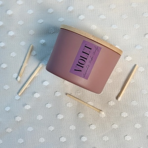 Może przedstawiać: Fioletowa świeca z drewnianą pokrywką i etykietą z napisem "VIOLET Lavender Tonka Woody". Kilka zapałek jest rozrzuconych wok&oacute;ł świecy na białej, kropkowanej powierzchni. Świeca ma kształt cylindryczny.