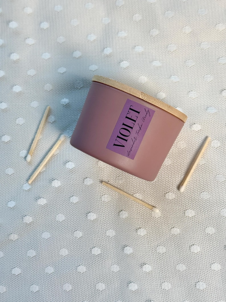 Może przedstawiać: Fioletowa świeca z drewnianą pokrywką i etykietą z napisem "VIOLET Lavender Tonka Woody". Kilka zapałek jest rozrzuconych wok&oacute;ł świecy na białej, kropkowanej powierzchni. Świeca ma kształt cylindryczny.