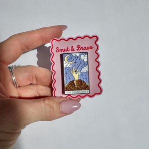 May include: A rectangular enamel pin featuring a tarot card design with a hand holding a needle and thread, set against a purple starry sky with a crescent moon and clouds. The text reads Smut & Bravo and The Seamstress on a pink backing.