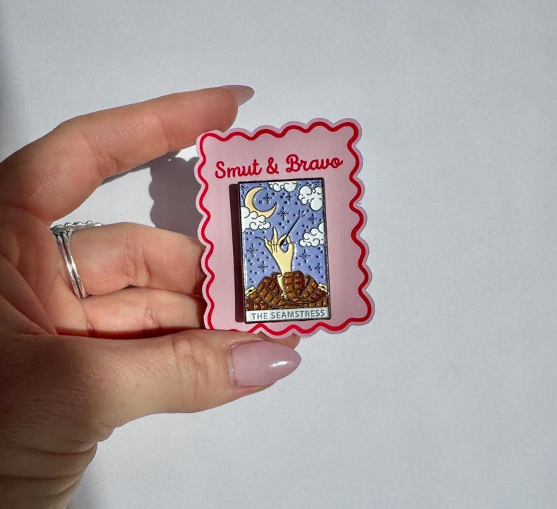 May include: A rectangular enamel pin featuring a tarot card design with a hand holding a needle and thread, set against a purple starry sky with a crescent moon and clouds. The text reads Smut & Bravo and The Seamstress on a pink backing.