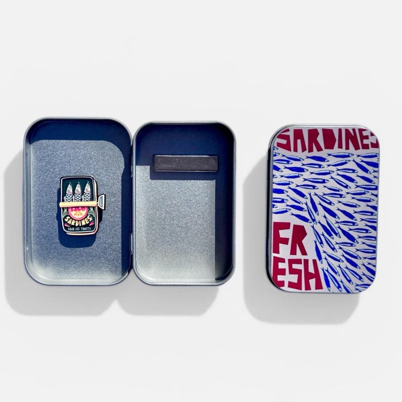 May include: A rectangular metal tin with a hinged lid featuring a blue and white sardine pattern and the red text SARDINES FRESH. Inside the open tin is a small enamel pin shaped like a sardine can with three fish and the text SARDINES POUR LES TOASTS.