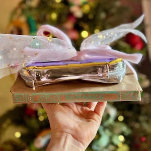 Może przedstawiać: Srebrna kosmetyczka z fioletowym i żółtym wykończeniem, owinięta różową kokardą z tiulu z błyszczącymi złotymi gwiazdkami. Kosmetyczka leży na brązowej książce z napisem "remarkable" na okładce.