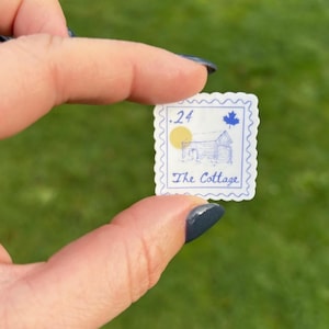 K&ouml;nnte beinhalten: Ein kleiner, quadratischer Aufkleber mit blauem und wei&szlig;em Design. Der Aufkleber zeigt eine Zeichnung eines Ferienhauses, ein Ahornblatt und den Text ".24 The Cottage". Der Hintergrund ist ein verschwommenes Gr&uuml;n.