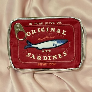 May include: A red cosmetic bag shaped like a sardine can, with the words "ORIGINAL SARDINES" and a fish illustration. The bag has a silver zipper and trim, and the text "IN PURE OLIVE OIL" is visible. Net weight is 3.75 oz.
