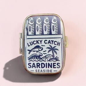 May include: A rectangular metal tin with a hinged lid featuring a dark blue and white graphic of four sardines, a fish, a palm tree, and ocean waves. The text reads LUCKY CATCH SARDINES SEASIDE. The container has a metallic gold-toned frame.