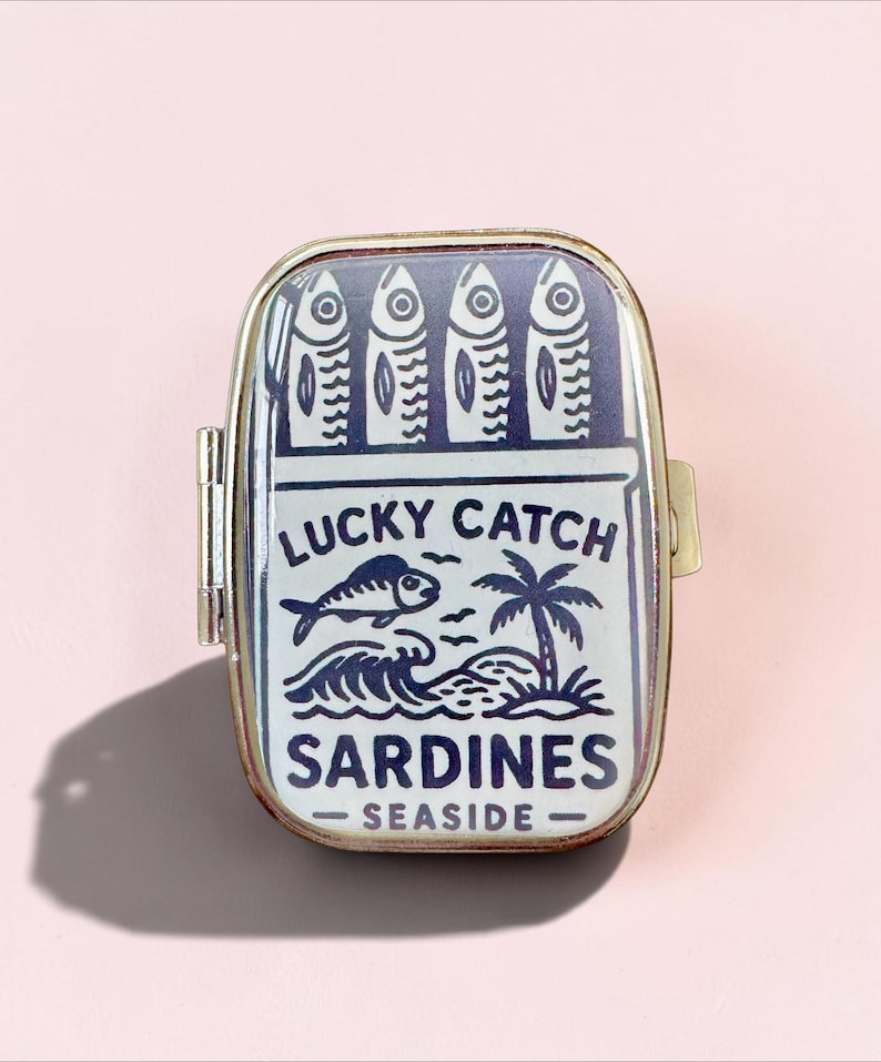 May include: A rectangular metal tin with a hinged lid featuring a dark blue and white graphic of four sardines, a fish, a palm tree, and ocean waves. The text reads LUCKY CATCH SARDINES SEASIDE. The container has a metallic gold-toned frame.