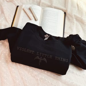 Puede incluir: Una sudadera negra de cuello redondo con el texto "VIOLENT LITTLE THING" y un gr&aacute;fico de murci&eacute;lago. La sudadera est&aacute; sobre una tela blanca con lunares, con un libro abierto y dos l&aacute;pices de color claro en el fondo.