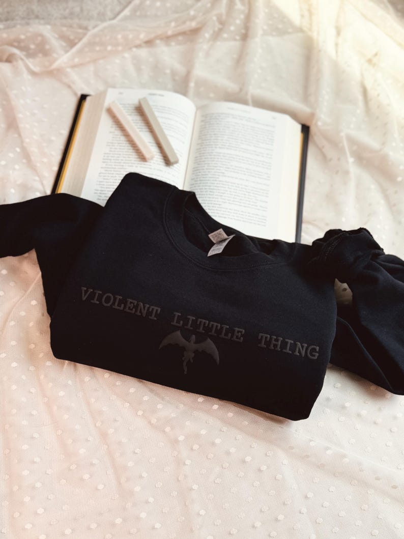 Puede incluir: Una sudadera negra de cuello redondo con el texto "VIOLENT LITTLE THING" y un gr&aacute;fico de murci&eacute;lago. La sudadera est&aacute; sobre una tela blanca con lunares, con un libro abierto y dos l&aacute;pices de color claro en el fondo.