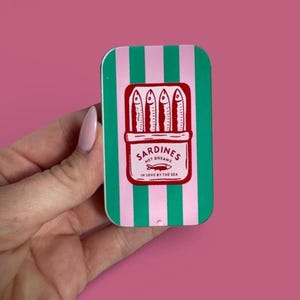 May include: A rectangular metal tin featuring vertical green and pink stripes. The center displays a red graphic of a sardine can with four fish inside. The text reads SARDINES NET DREAMS IN LOVE BY THE SEA with a small fish icon below the text.