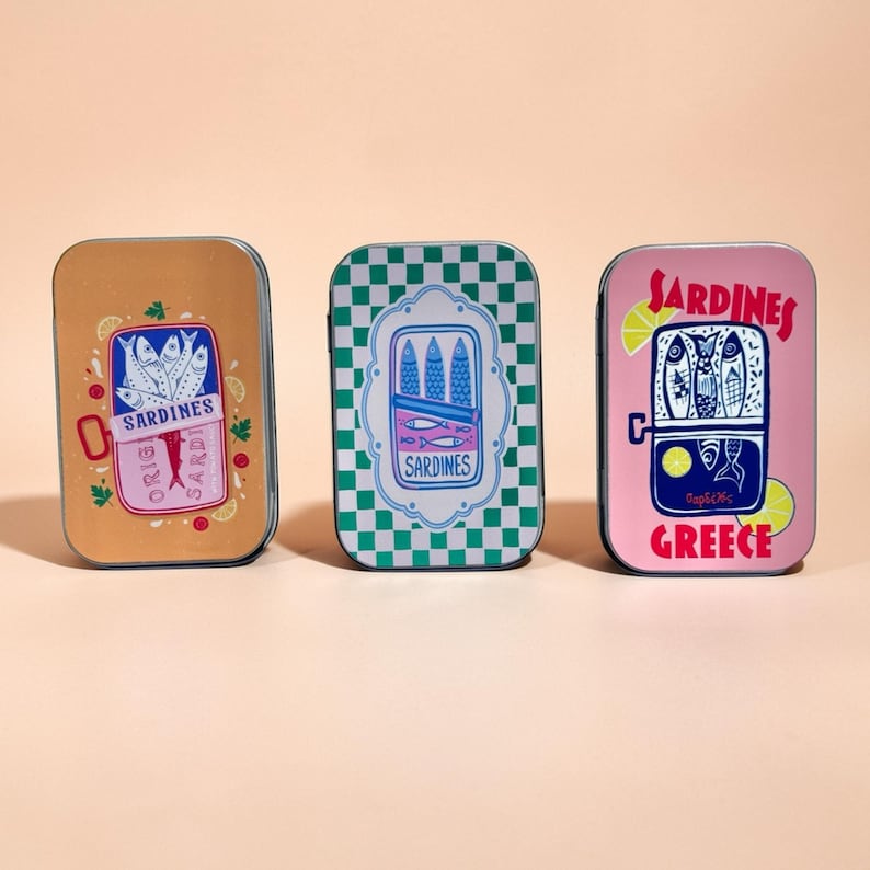May include: Three rectangular metal tins featuring sardine-themed illustrations. The left tin is tan with a sardine can graphic. The center tin has a green and white checkerboard pattern. The right tin is pink with the text SARDINES GREECE.
