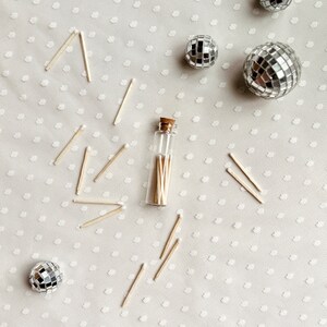 May include: A small glass bottle filled with wooden matches, surrounded by silver disco balls on a white background with a polka dot pattern.
