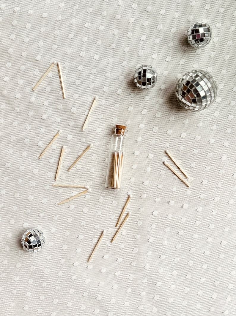May include: A small glass bottle filled with wooden matches, surrounded by silver disco balls on a white background with a polka dot pattern.