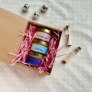 May include: A gift box filled with pink crinkle paper and three small candles in gold tins. The candles are labeled "Cassian", "Azriel", and "Rhysand". Two small glass vials with matches are also included.