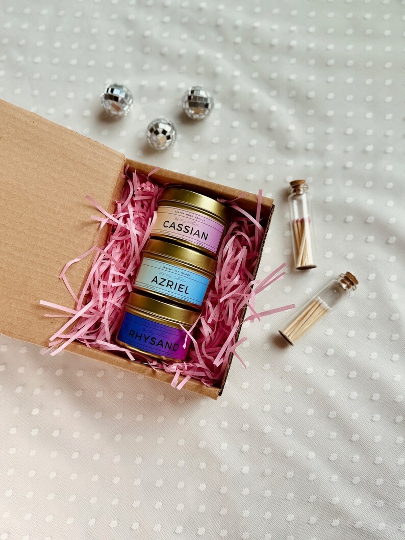 May include: A gift box filled with pink crinkle paper and three small candles in gold tins. The candles are labeled "Cassian", "Azriel", and "Rhysand". Two small glass vials with matches are also included.