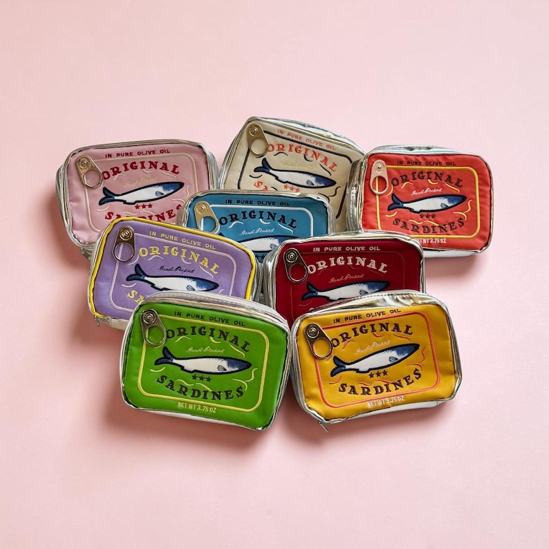 May include: A collection of rectangular zippered pouches designed to resemble sardine tins in various colors including pink, cream, blue, red, purple, green, and yellow. Each pouch features a sardine graphic, a metal pull tab, and text.