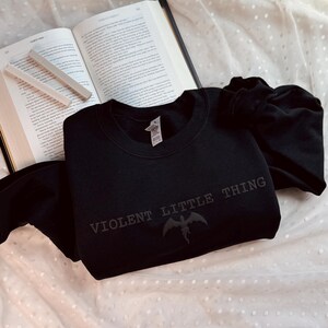 Puede incluir: Sudadera negra de cuello redondo con el texto "VIOLENT LITTLE THING" y un gr&aacute;fico de criatura alada. La sudadera est&aacute; sobre una superficie blanca con lunares, junto a un libro abierto y tiza.