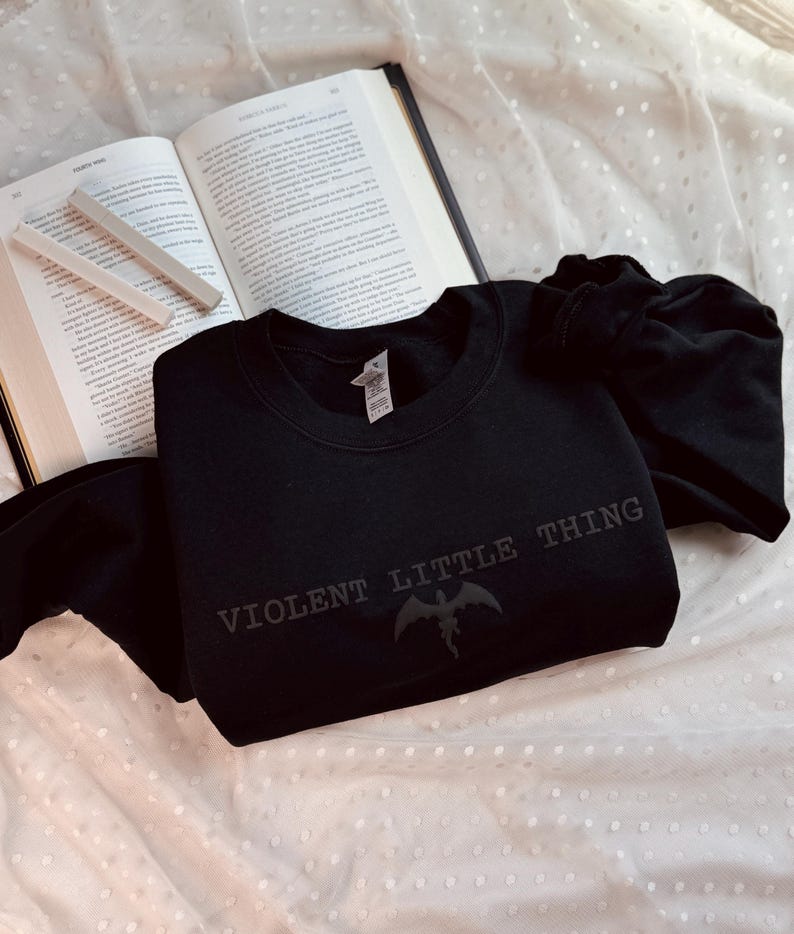 Puede incluir: Sudadera negra de cuello redondo con el texto "VIOLENT LITTLE THING" y un gr&aacute;fico de criatura alada. La sudadera est&aacute; sobre una superficie blanca con lunares, junto a un libro abierto y tiza.