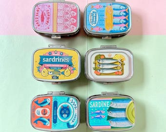Puede incluir: Seis latas metálicas rectangulares con esquinas redondeadas, cada una con diseños gráficos coloridos de sardinas, peces y gatos. Las etiquetas incluyen textos como Sardine Love Club, Totally Hooked, Sardrines with lemon oil y Sardine.