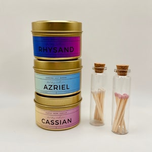 May include: Three gold candle tins with labels that read "Rhysand", "Azriel", and "Cassian". The candles are stacked on top of each other. Two small glass jars with cork tops contain matches.