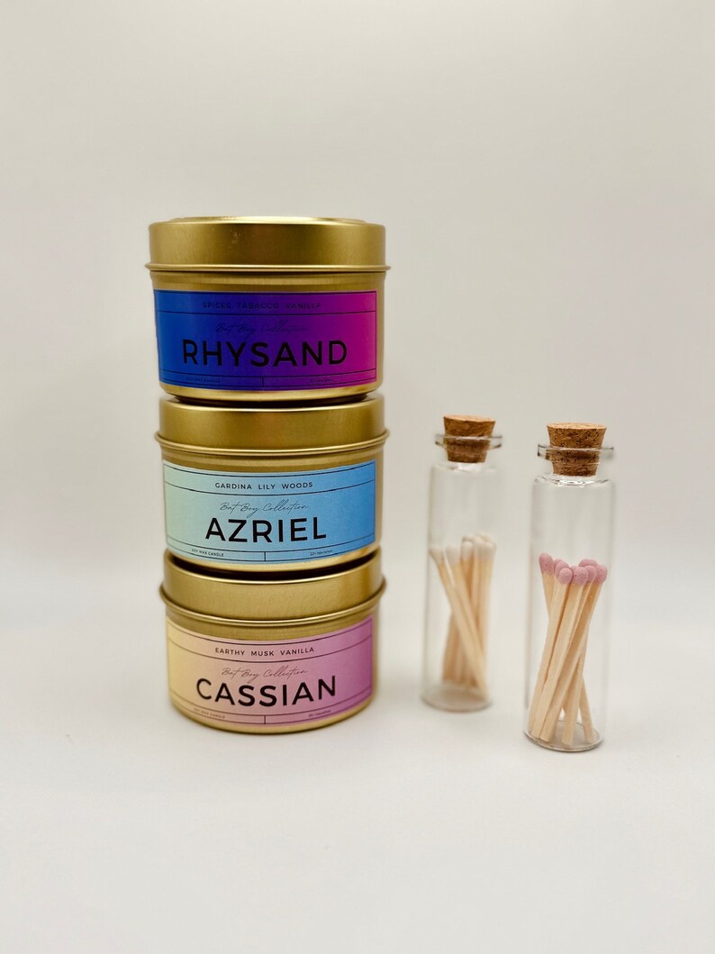 May include: Three gold candle tins with labels that read "Rhysand", "Azriel", and "Cassian". The candles are stacked on top of each other. Two small glass jars with cork tops contain matches.