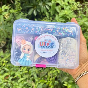 May include: A clear plastic box filled with sensory kit components. The kit includes blue and white glittery play dough, a doll figurine, and various pastel-colored decorative elements. The box has the text "Liliput tiny hands" on it.