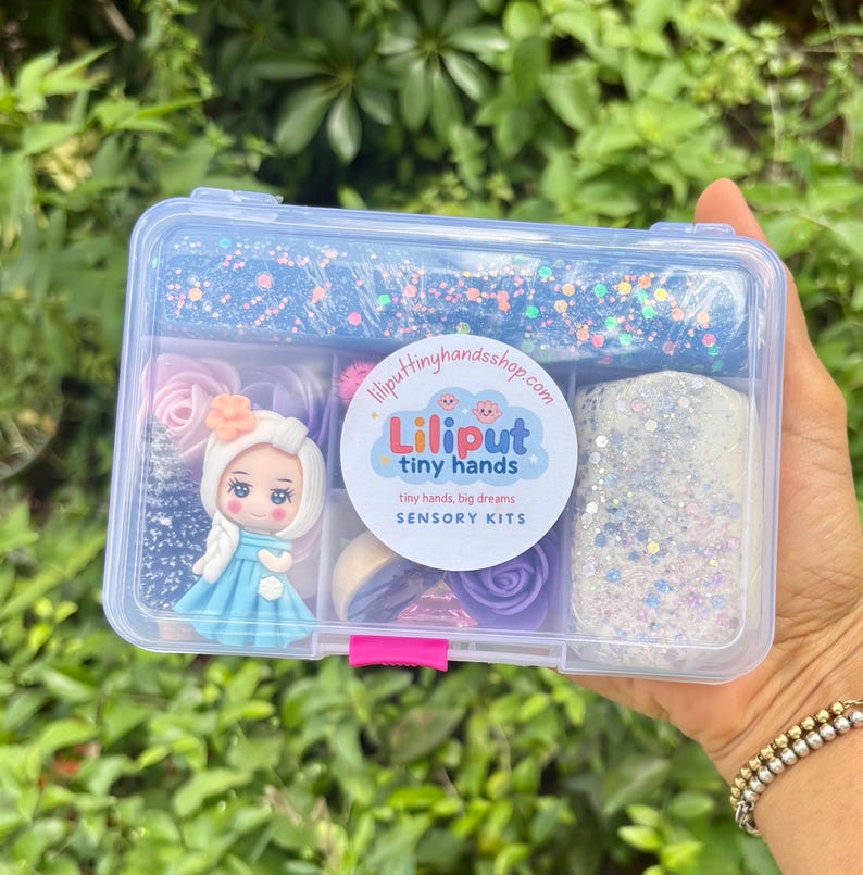 May include: A clear plastic box filled with sensory kit components. The kit includes blue and white glittery play dough, a doll figurine, and various pastel-colored decorative elements. The box has the text "Liliput tiny hands" on it.