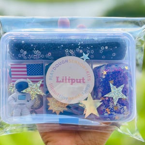 May include: A clear plastic container filled with various items for a space-themed playdough sensory kit. The kit includes a miniature astronaut figurine, an American flag, star-shaped glitter, and blue and purple playdough with glitter.