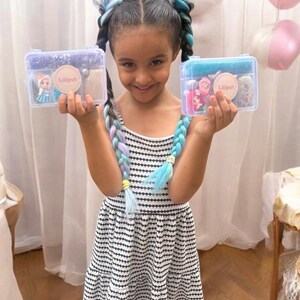 May include: A young girl smiles, holding two clear plastic boxes. Each box contains accessories and a sticker with the text "Liliput". The girl wears a black and white striped dress and has braided hair with blue, purple, and white extensions.