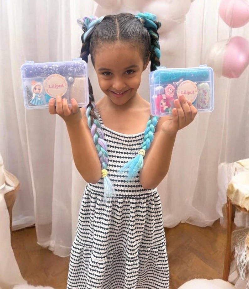 May include: A young girl smiles, holding two clear plastic boxes. Each box contains accessories and a sticker with the text "Liliput". The girl wears a black and white striped dress and has braided hair with blue, purple, and white extensions.