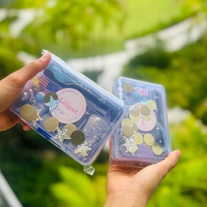 May include: Two clear plastic containers filled with colourful craft supplies, including glitter, stars and circles. The containers are labelled "Liliput Playdough Shop".