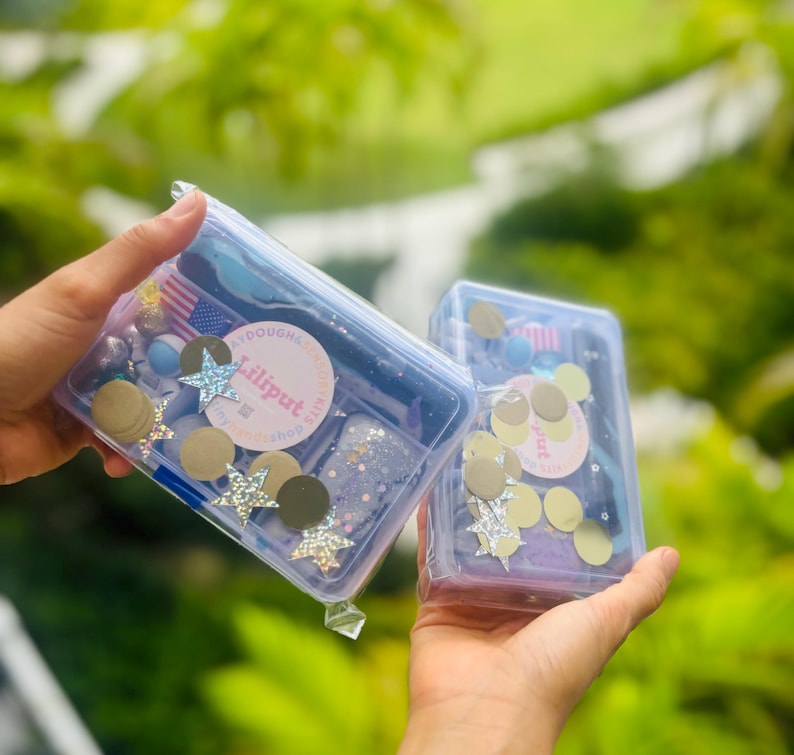 May include: Two clear plastic containers filled with colourful craft supplies, including glitter, stars and circles. The containers are labelled "Liliput Playdough Shop".