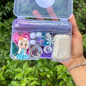 May include: A clear plastic box filled with various craft supplies. The box contains glittery purple slime, a miniature doll in a blue dress, pink roses, white pom-poms, and decorative gems. The theme appears to be inspired by a popular animated movie.
