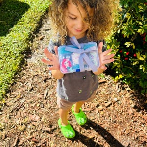 May include: A clear plastic box tied with a light blue ribbon, containing various craft supplies. The box features a sticker of a cartoon mermaid and the name "Lili". The box is held by a person wearing a gray t-shirt and green clogs.