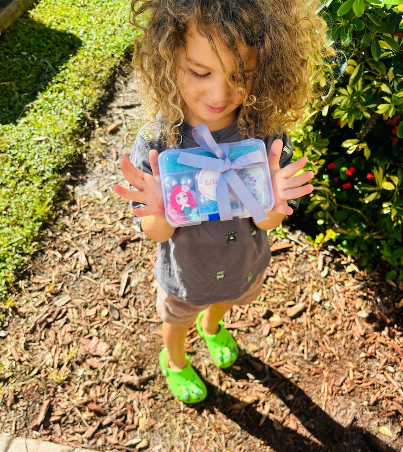 May include: A clear plastic box tied with a light blue ribbon, containing various craft supplies. The box features a sticker of a cartoon mermaid and the name "Lili". The box is held by a person wearing a gray t-shirt and green clogs.