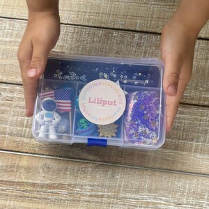 May include: A clear plastic container with a blue lid filled with various items for a space-themed play dough sensory kit. The kit includes a small astronaut figurine, a blue alien, a star-shaped container with glitter, a small American flag, a purple play dough with blue and silver glitter, and a round sticker with the text "Lilliputillputtinyhands.shop".
