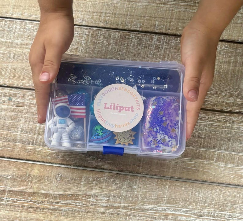 May include: A clear plastic container with a blue lid filled with various items for a space-themed play dough sensory kit. The kit includes a small astronaut figurine, a blue alien, a star-shaped container with glitter, a small American flag, a purple play dough with blue and silver glitter, and a round sticker with the text "Lilliputillputtinyhands.shop".