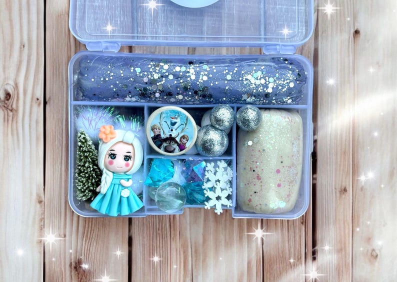 May include: A clear plastic box filled with various craft supplies. The box contains glittery blue slime, a doll, silver balls, and other decorative items. The theme appears to be inspired by the movie "Frozen".