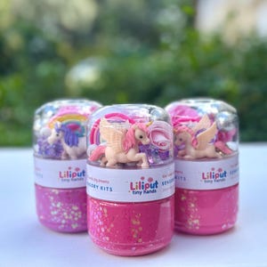 Pode incluir: Três kits sensoriais com fundos rosa brilhantes e tampas transparentes. Cada kit contém uma figura de unicórnio, um arco-íris e flores. Os kits têm um rótulo que diz "Liliput tiny hands". O fundo é verde desfocado.
